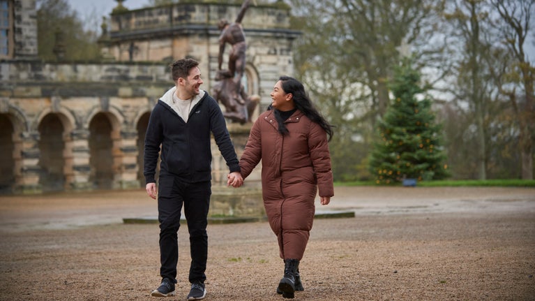 A couple holding hands in winter clothes walking with part of the building and statue in the background